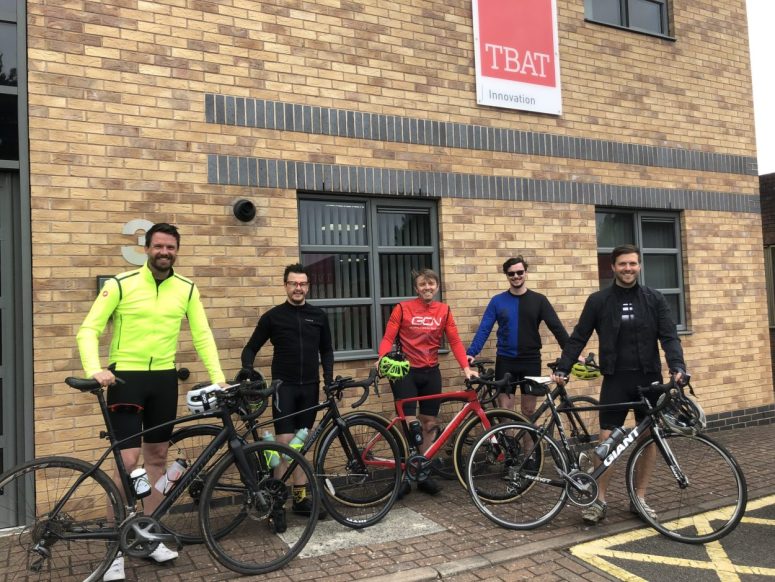 5 cyclists stood with their bikes outside of TBAT Innovation Ltd office building, with TBAT logo above them